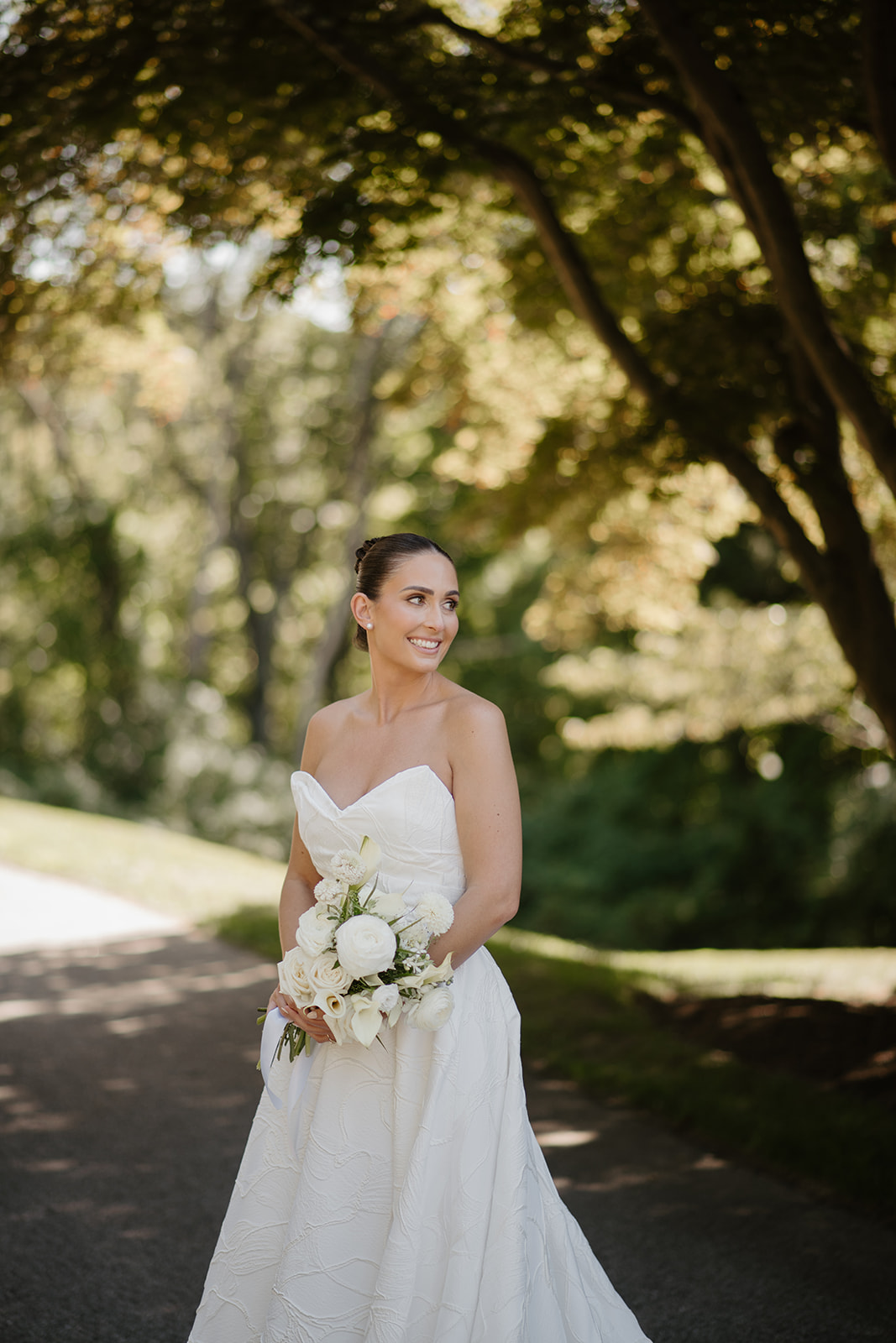 Radiant bride smiling warmly while holding her white bouquet under the shade of trees.
